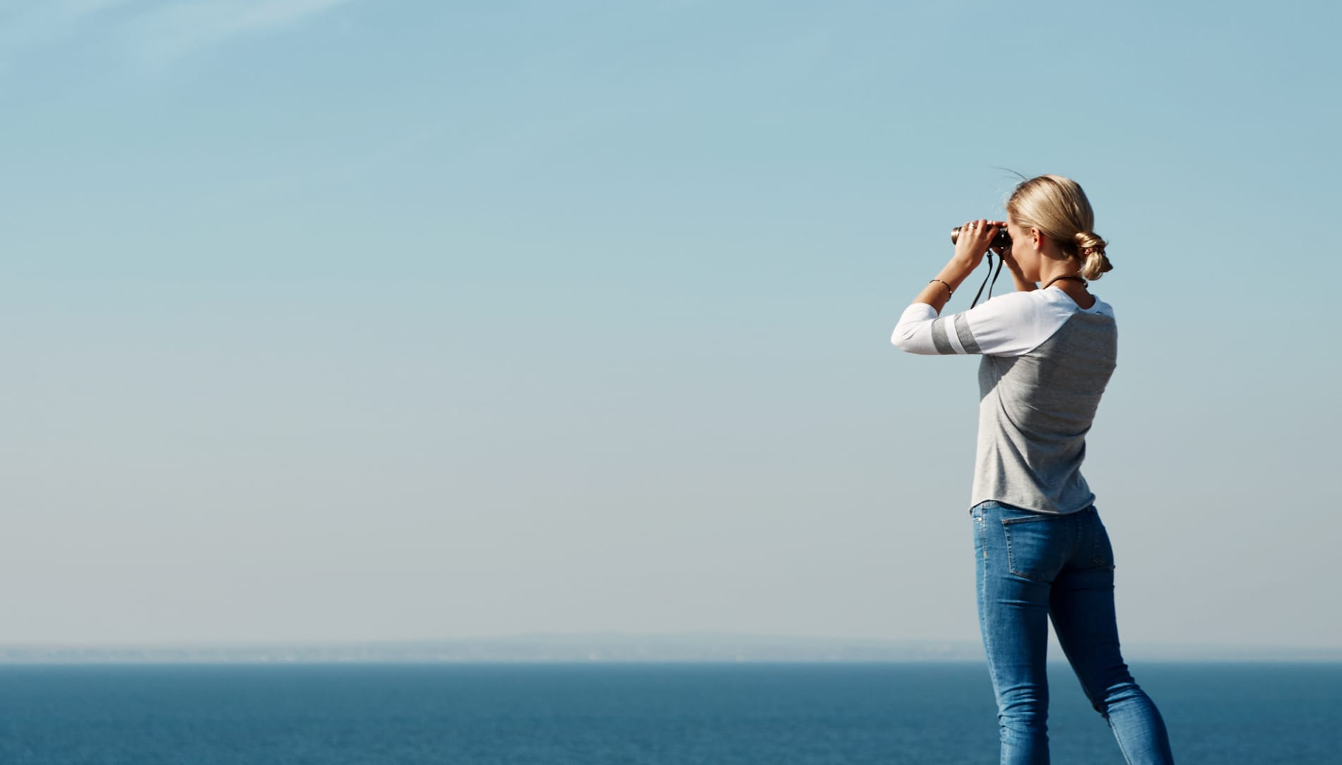 Eine junge Dame schaut mit einem Fernglas auf das offene Meer.